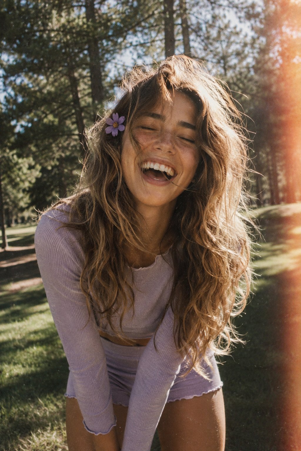 A candid, film-style portrait of a joyful young woman with long, voluminous, wavy light brown hair with blonde highlights, laughing heartily with her eyes closed and mouth open in a genuine, ecstatic expression. She has a small purple flower tucked behind her ear, and her hair is slightly windswept across her face. She is leaning slightly forward toward the camera, wearing a matching lavender-colored long-sleeve top and shorts set. The background is a sun-drenched outdoor park with green grass, shaded areas, and tall pine trees under bright sunlight, captured with a vintage 35mm film aesthetic, subtle grain, warm sun flares, and a soft, desaturated color palette.